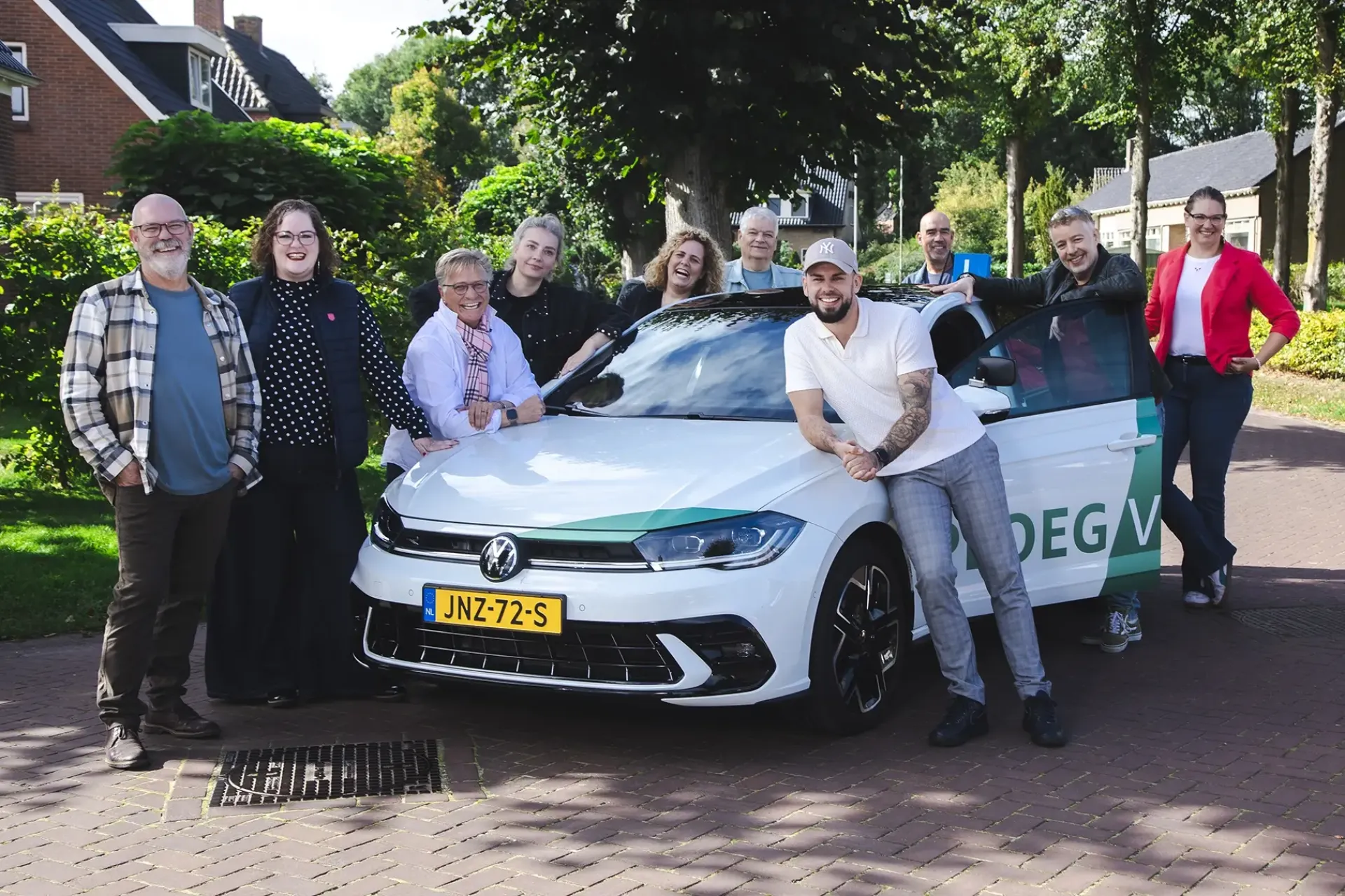 Een groep mensen poseert rond een lesauto op een zonnige dag in een woonwijk.